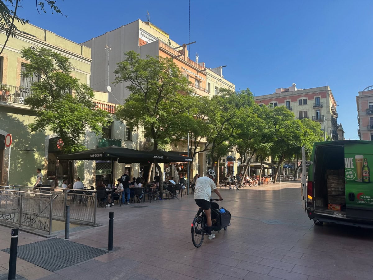 A tiled road in Barcelona with car, person riding a bicycle, trees, and outdoor dining area of a cafe.