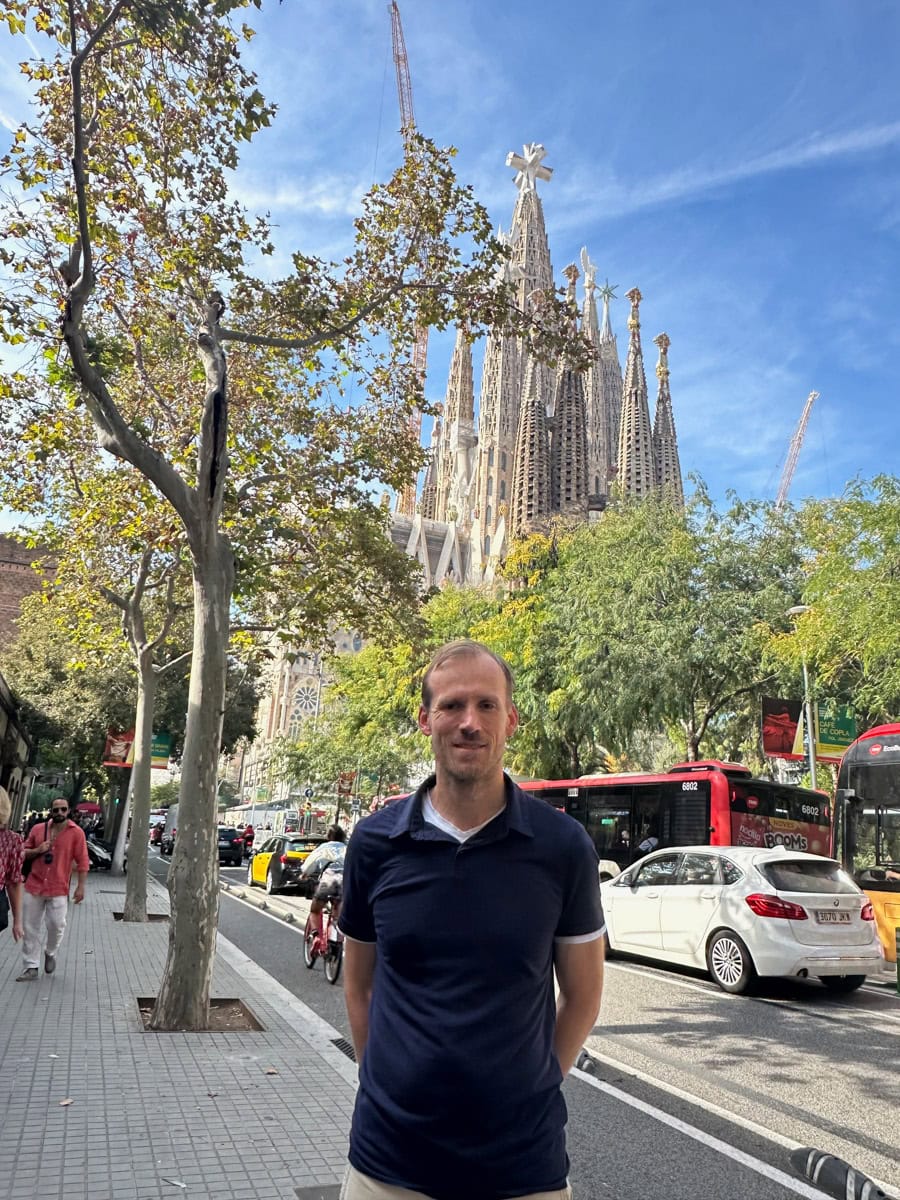 A man standing on a sidewalk with the road with cars, trees, and the Sagrada Familia church in the background.