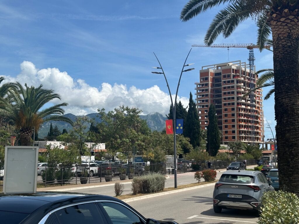 View of street in Budva lined with palm trees and large residential block of flats under construction. There is a crane next to the building.