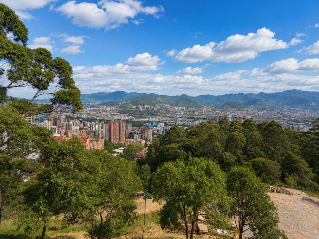 View from a hill surrounding Medellin of a city of residential buildings and lots of green and mountains in the back