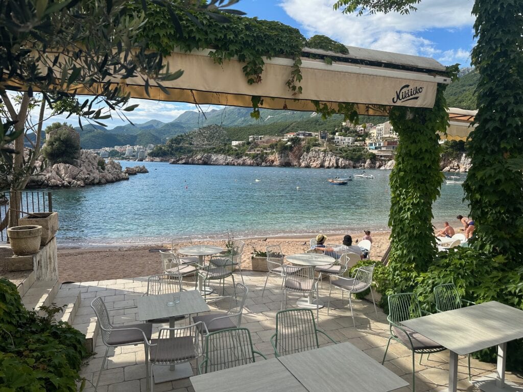 Gorgeous view of a beach, sea and surrounding green hills, and in front a few tables and chairs of a local restaurant or cafe, near Budva, Montenegro