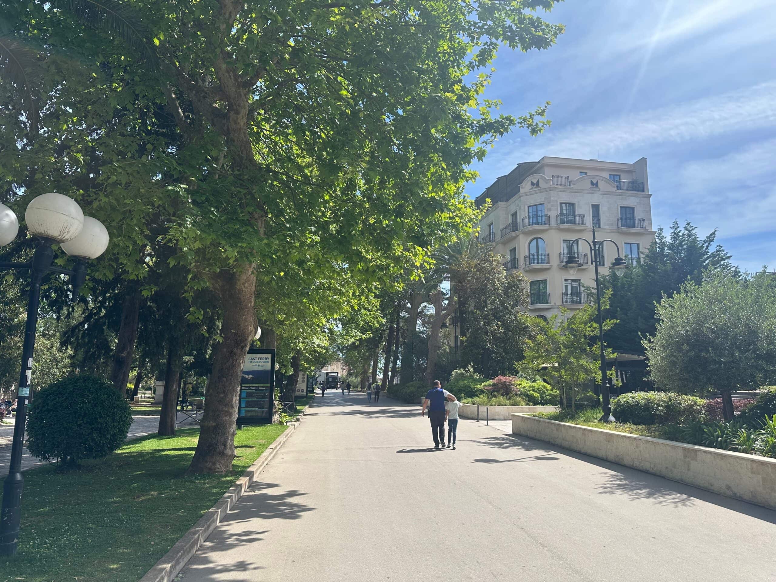 Pedestrian broad path lined with trees and a classical residential building/hotel style building in the distance in central Budva, Montenegro