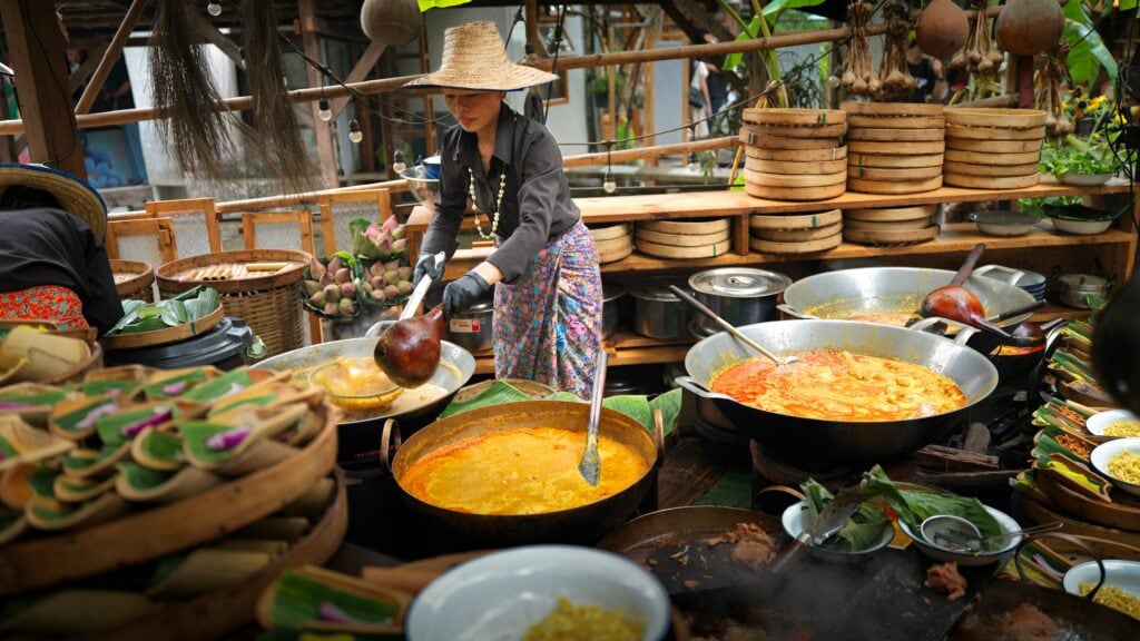 A local Thai woman cooking in an open kitchen with big woks, all flled with colourful soups and food, in Chiang Mai, Thailand
