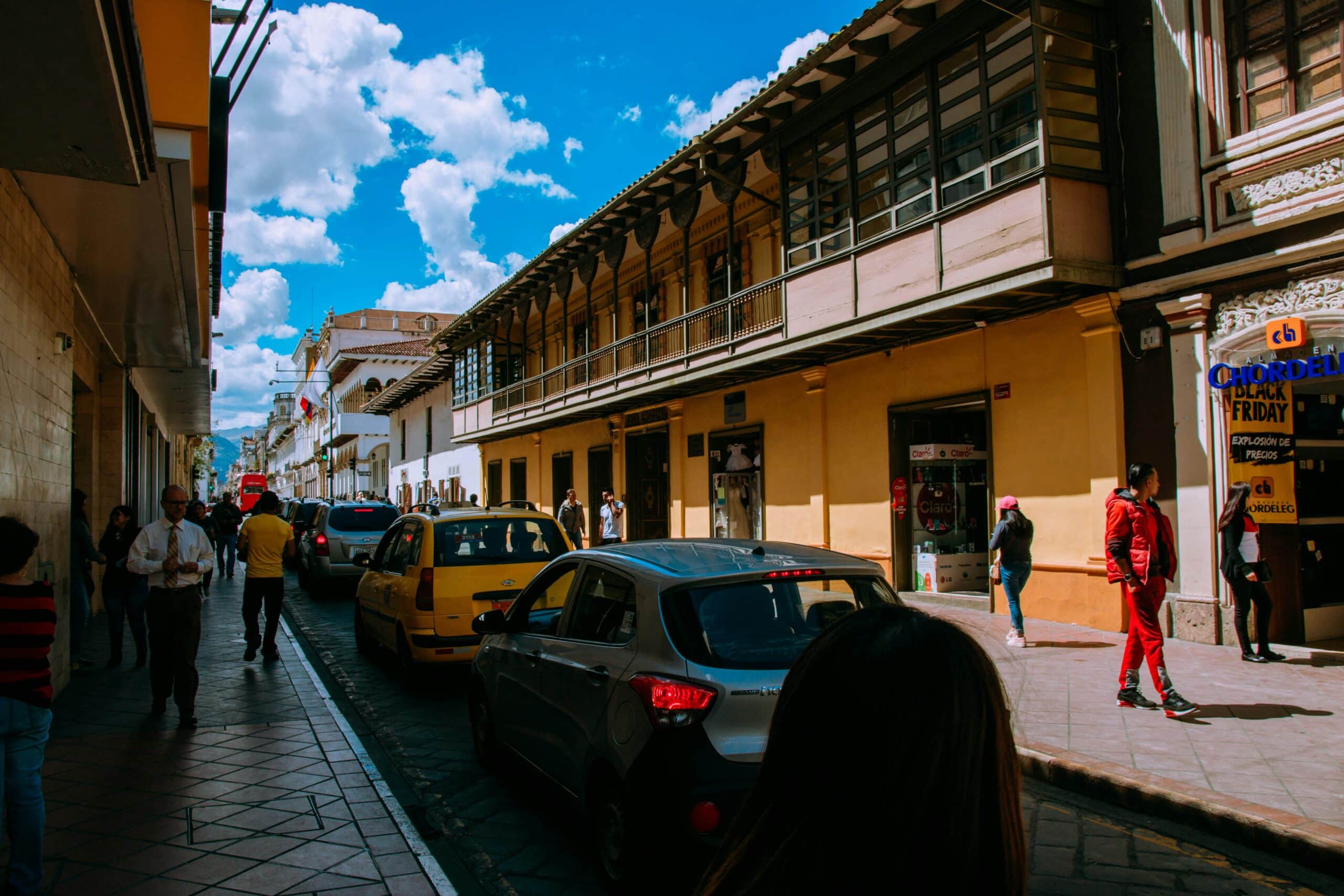 A street in central Cuenca, Ecuador. Cars are lining up and people walking on the sidewalk next to shops